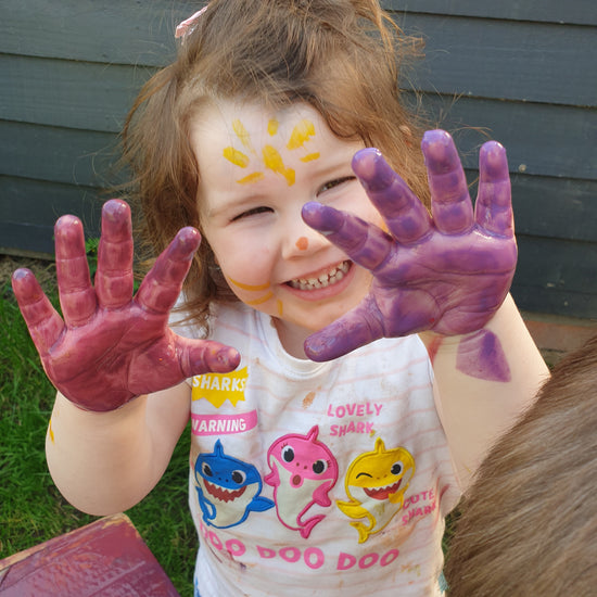 Playful child with purple and pink painted hands  sharing the joy of connecting with play,  building mindfulness through play using all the senses and been in sync with nature